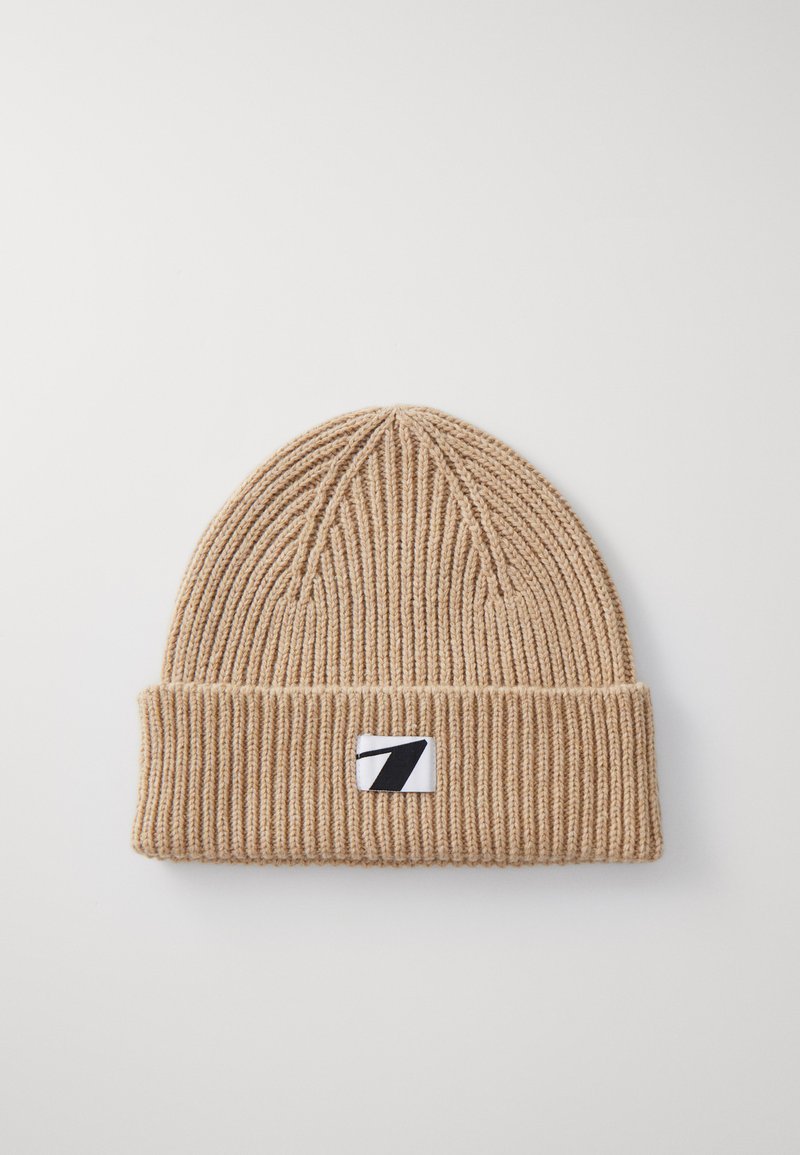Beige gebreide muts met een geribbelde textuur, voorzien van een omgevouwen rand en een klein zwart-wit logo op de voorkant.