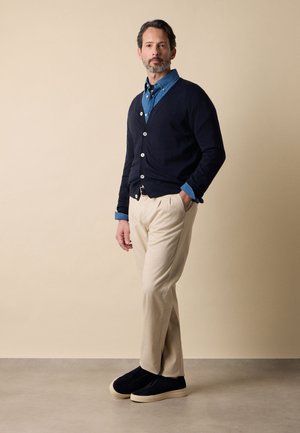 Mann steht mit Händen in den Taschen, trägt eine marineblaue Strickjacke, ein blaues Hemd mit Knöpfen, beige Hose und schwarze Slipper vor beigem Hintergrund.
