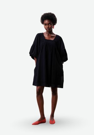 Femme aux cheveux courts et bouclés portant une robe noire ample à manches bouffantes et des ballerines rouges, debout devant un fond uni.