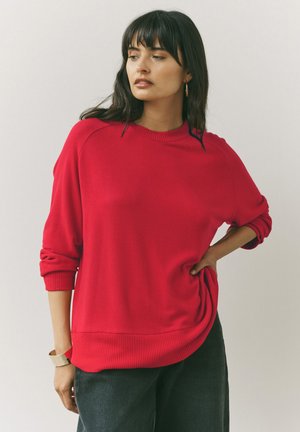 Crvena pleta sweater sa rebrastim okovratnikom i dugim rukavima. Ima opušteni kroj i rebraste detalje na rubu, uparen s tamnim hlačama.