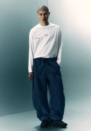 Ung mand med barberet hoved iført hvid langærmet skjorte og løse marineblå bukser, stående i et blødt oplyst minimalistisk rum.