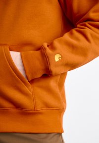 Orange hoodie made of soft fabric, featuring a front pocket and a small golden logo on the sleeve. Smooth texture and relaxed fit.