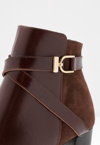 Bottine marron combinant cuir brillant et daim, avec un détail de boucle dorée et des surpiqûres contrastantes.