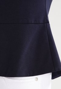 Mörkblå peplumtop i ett slätt tyg, med en utsvängd nederdel och sömlös design, kombinerad med vita byxor och en silverknappdetalj.