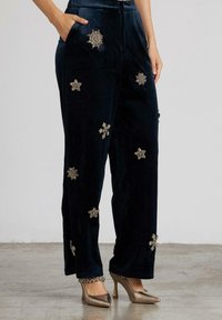 Pantalones de terciopelo azul marino con embellishments metálicos en forma de estrella, bolsillos laterales y un corte relajado, combinados con tacones beige de punta afilada.