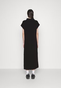 Schwarzes gestricktes Kleid mit kurzen Ärmeln, hohem Halsausschnitt und gerader, langer Silhouette, kombiniert mit klobigen schwarzen Schuhen und weißen Socken.