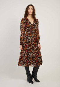 Vrouw in een mouwloze V-hals midi-jurk met oranje, rode en beige bladeren en dierenprints, gecombineerd met zwarte kniehoge laarzen.
