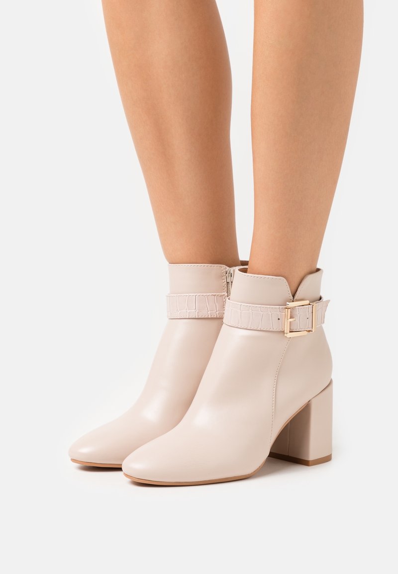Bottes en cuir beige montant jusqu'à la cheville, avec talons carrés, brides décoratives à motif crocodile et boucles dorées, portées aux jambes.