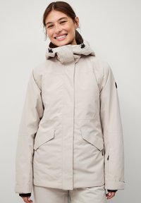 Lys beige udendørsjakke med en tætsiddende hætte, kontrasterende sorte hardware, lynlåslommer og lynlåse ved manchetterne. Glat tekstur og skræddersyet design.