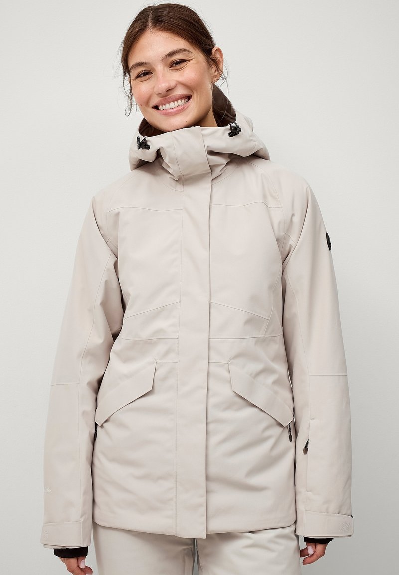 Lys beige udendørsjakke med en tætsiddende hætte, kontrasterende sorte hardware, lynlåslommer og lynlåse ved manchetterne. Glat tekstur og skræddersyet design.