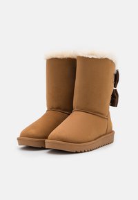 Bottes en suède tan avec un design haut, doublure en fausse fourrure blanche et un nœud décoratif marron sur le côté. Semelle en caoutchouc texturée pour une meilleure adhérence.