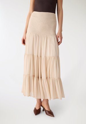 Donna che indossa una maxi gonna beige a balze con vita arricciata, abbinata a mules con tacco marroni e punta appuntita su sfondo bianco.