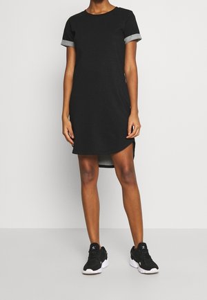 Woman wearing a short-sleeve black dress with rolled cuffs and black sneakers with white soles, standing against a plain background.