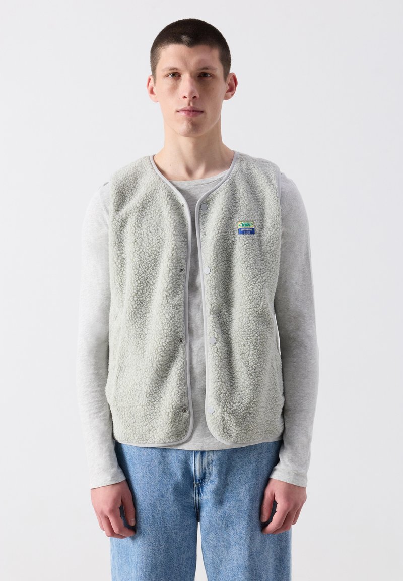 Jauns vīrietis, valkājot gaiši pelēku vilnas vestīti pār garām piedurknēm krekla un zilas džinsas, stāv pret vienkāršu baltu fona.