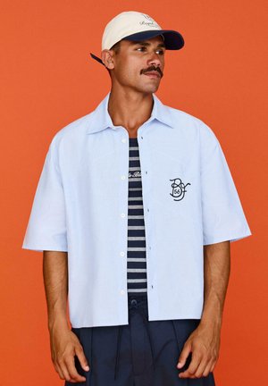 Mand iført en hvid kasket, lyseblå kortærmet skjorte med knapper over en sort og hvidstribet skjorte, stående mod en orange baggrund.