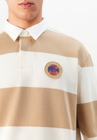 Vyriškis, vilkintis kreminę ir baltą juostuotą polo marškinėlį su baltu apykakle ir apvaliu pleistru, kuriame užrašyta "Tennis Heritage" su mėlynu krokodilo logotipu.