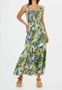 Robe maxi avec motif de feuilles vertes et bleues, corsage froncé, encolure carrée et ourlet à volants. Portée avec des sandales compensées couleur sable.