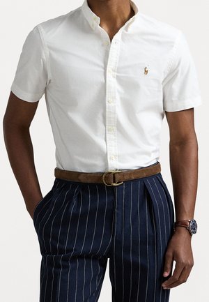 Man draagt een wit overhemd met korte mouwen en een klein logo, marineblauwe pantalon met fijne strepen, een bruine suède riem en een horloge met een bruine leren band.