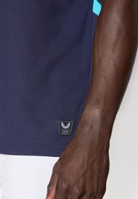 Chemise texturée bleu marine avec un petit logo de marque noir et blanc, présentant des accents bleu clair sur les côtés et un design en tissu côtelé.
