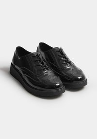 Chaussures brogues en cuir verni noir avec des détails en pointe et des semelles en caoutchouc noir, présentées côte à côte sur un fond blanc.