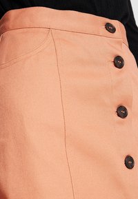 Jupe en denim couleur pêche avec une taille ajustée, comportant cinq boutons noirs sur le devant et des coutures diagonales subtiles pour la texture.