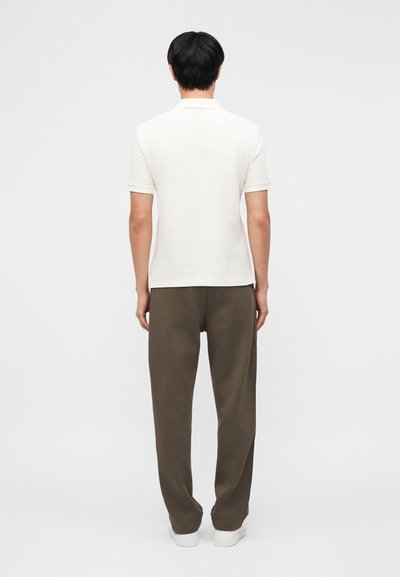 Άνδρας που στέκεται γυρισμένος με την πλάτη φοράει λευκό πόλο με κοντά μανίκια, καφέ παντελόνι και λευκά αθλητικά παπούτσια μπροστά σε απλό φόντο.