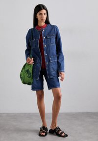 Dżinsowa kurtka z złotymi guzikami i naszywanymi kieszeniami, w połączeniu z dżinsowymi szortami. Zestaw dopełnia zielona torebka o teksturowanej powierzchni i czarne sandały.