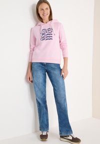 Sweat à capuche rose avec texte graphique bleu marine, associé à un jean bleu clair. Des baskets marron avec des accents blancs complètent la tenue.