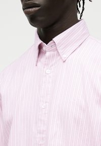 Κοντινό πλάνο ενός ατόμου που φοράει ένα ροζ και λευκό κάθετα ριγέ πουκάμισο με γιακά με κουμπιά και κεντημένο λογότυπο στο στήθος.