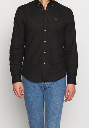 Mężczyzna ubrany w czarną koszulę z długim rękawem zapinaną na guziki i jasnoniebieskie jeansy, stojący na jednolitym tle.