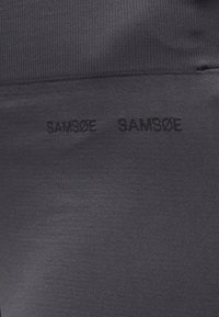 Le tissu noir extensible à texture lisse présente le nom de la marque "SAMSOE" en impression sombre et subtile répétée le long de la ceinture.