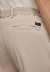 Pantalon beige en tissu léger, avec une poche arrière unique à ouverture horizontale et une étiquette de marque. Texture lisse.