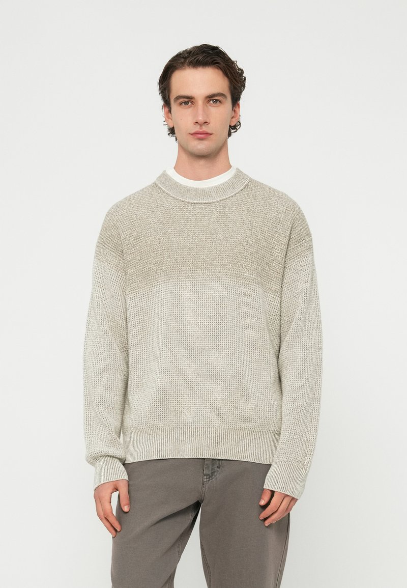 Ανοιχτό γκρι πλεκτό πουλόβερ με υφή και σχέδιο, ριμπ μανσέτες και ψηλό γιακά. Φορεμένο πάνω από λευκό πουκάμισο. Συνδυασμένο με γκρι παντελόνι.