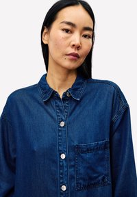 Denim overhemd in donkerblauw met een kraag, knopen aan de voorkant en een borstzak. Heeft een ontspannen pasvorm en een gladde textuur.