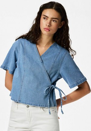 Ung kvinde med krøllet mørkt hår iført en kortærmet, omslagsstyle denimtop bundet ved taljen og hvide bukser, poserer med hånden på hoften.