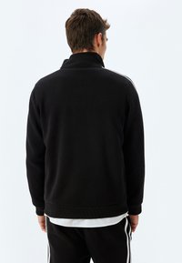 Sudadera negra con cremallera y cuello alto, con puños y dobladillo acanalados, que presenta rayas blancas en los laterales y una textura suave, llevada sobre pantalones oscuros.
