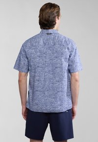 Camisa de manga corta en azul con un diseño texturizado y estampado. Con un dobladillo recto y un cuello trasero abotonado. Combinada con pantalones cortos oscuros.