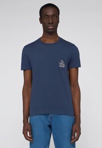 Camiseta de algodón azul marino con cuello redondo, mangas cortas y un bolsillo en el pecho con un detalle de logo. Llevada con jeans de color azul claro.