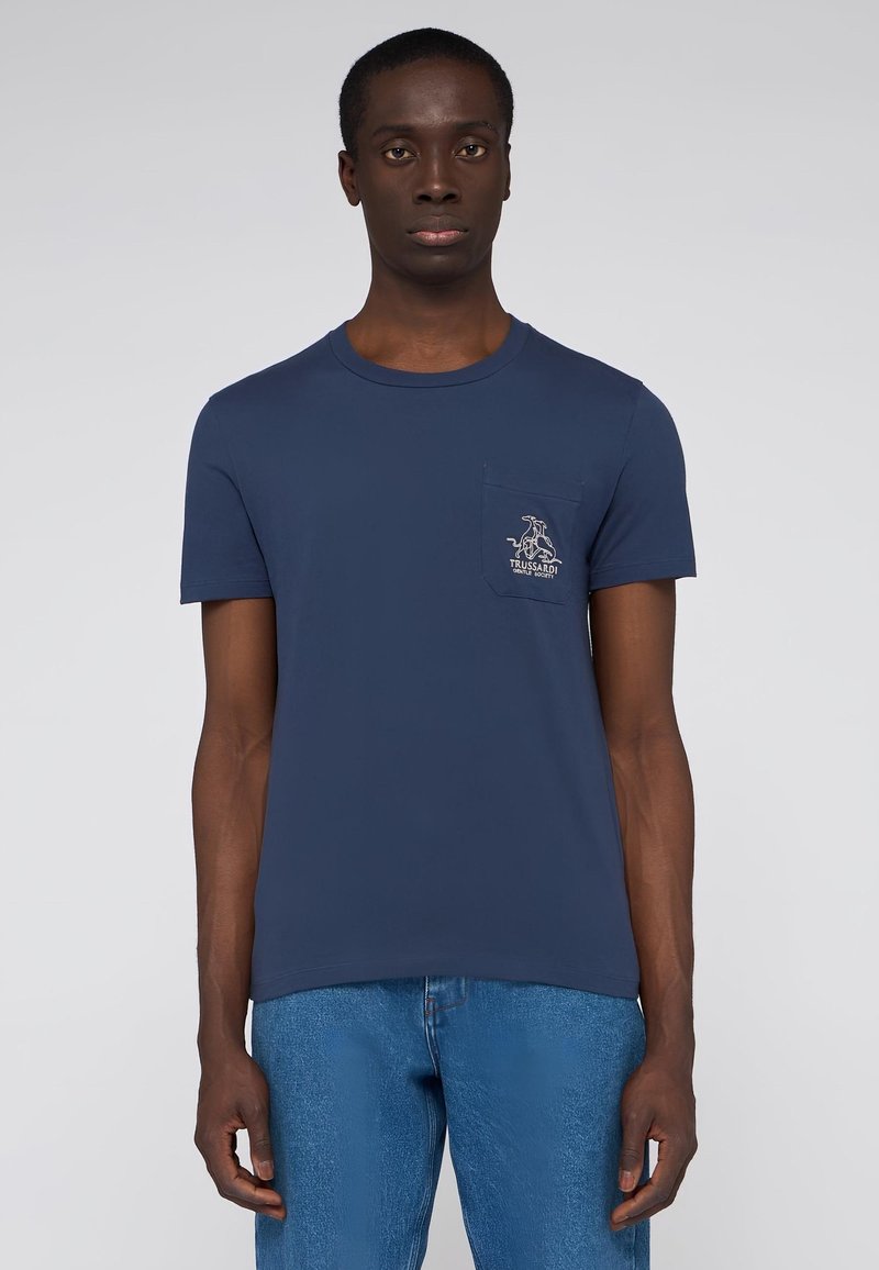 Camiseta de algodón azul marino con cuello redondo, mangas cortas y un bolsillo en el pecho con un detalle de logo. Llevada con jeans de color azul claro.