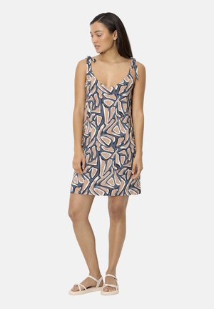 Mujer de pie con un vestido sin mangas y escote en V con patrones abstractos en beige y azul, combinado con sandalias blancas planas.