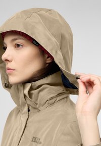 Giacca impermeabile beige con cappuccio, dotata di tasca con zip, elementi regolabili e fodera interna di colore blu scuro.