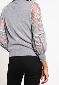 Gray pullover sweater with elasticized striped balloon sleeves and floral lace details on the shoulders, paired with black pants.
