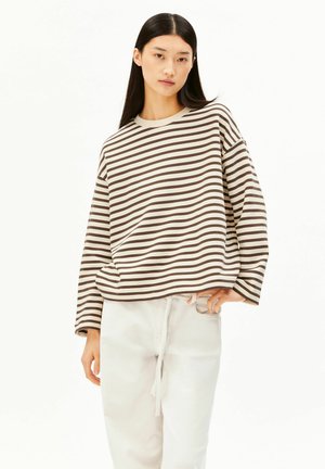 Giovane donna con lunghi capelli neri, indossa una camicia a maniche lunghe marrone e bianca a righe morbida e pantaloni bianchi con coulisse, in piedi su sfondo bianco.