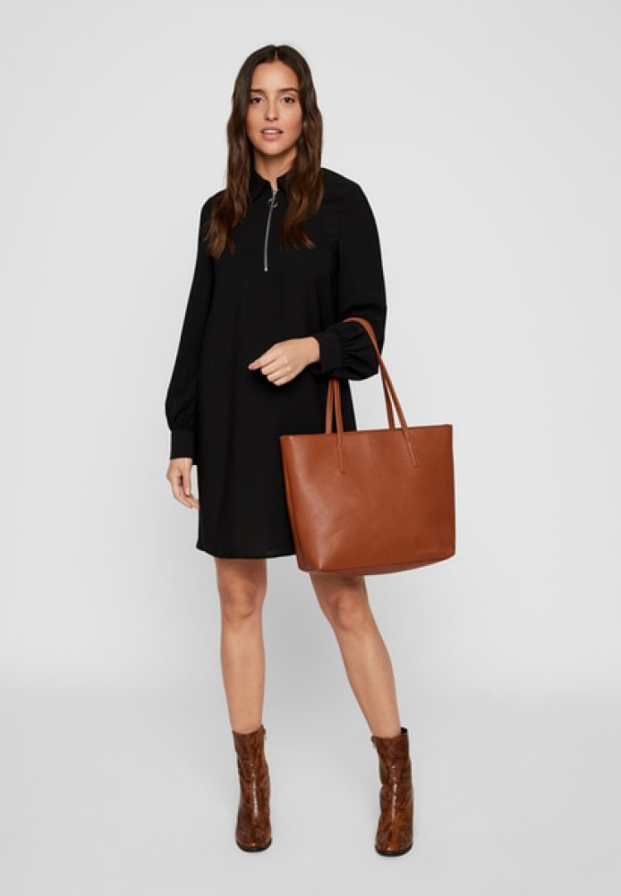 Vestido negro de manga larga con cuello de cremallera, acompañado de un bolso tote de cuero marrón. La modelo lleva botines de tacón marrones.