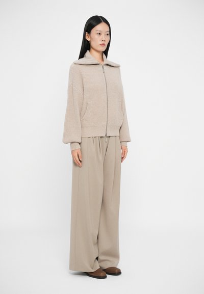 Beige zip-up genser med stor krage og to framlommer, kombinert med vide beige bukser og brune sko, på en nøytral bakgrunn.