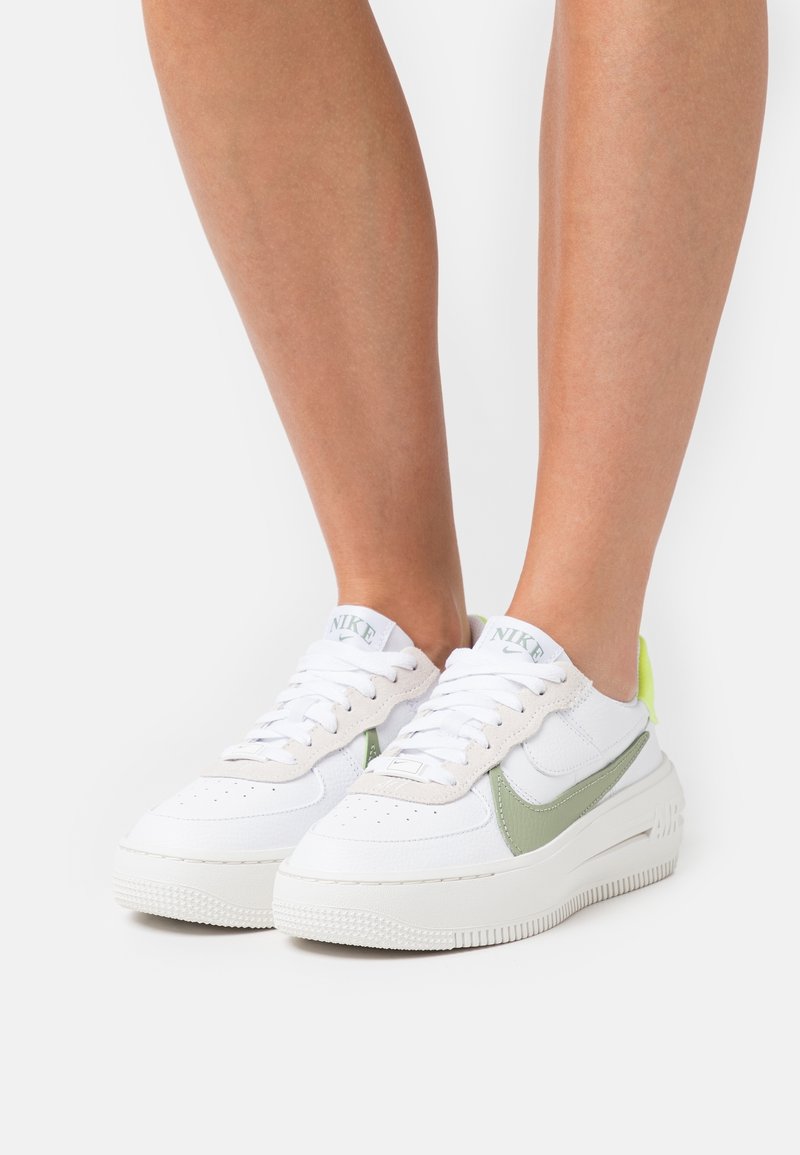 Personne portant des baskets Nike blanches et vert clair avec des lacets blancs, montrée de la moitié du mollet vers le bas sur un fond blanc uni.