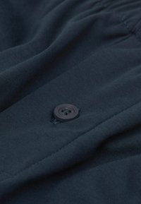 Ciemnonawowy materiał o gładkiej teksturze z widocznymi guzikami i przeszyciami, podkreślający miękki, zwiewny krój.