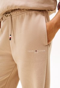 béžové tepláky Tommy Hilfiger s predným vreckom, pásom na šnúrku a logom na stehne, ruka odpočívajúca vo vrecku.