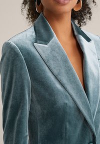 Sammetssak i turkos med krossad slagen, djup V-hals och texturerad tyg. Inkluderar två sidofickor och mjuka sömdetaljer.