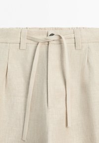 Beige linen pants with belt loops, a button closure, and a fabric tie belt at the waist in a casual style.